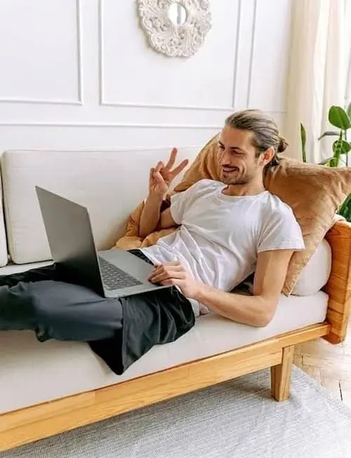 Zufriedener Chat-Moderator mit Kopfhörern arbeitet entspannt mit dem Laptop auf dem Sofa